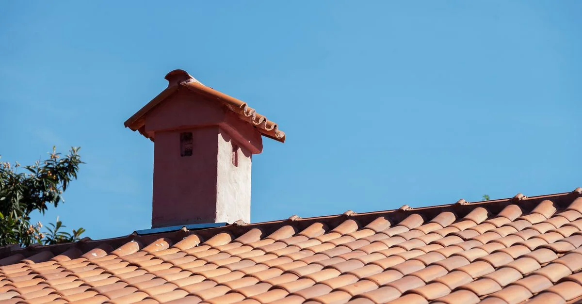 Primer plano de un tejado de tejas rojas y una chimenea bajo un cielo azul brillante, que muestra detalles arquitectónicos vibrantes.